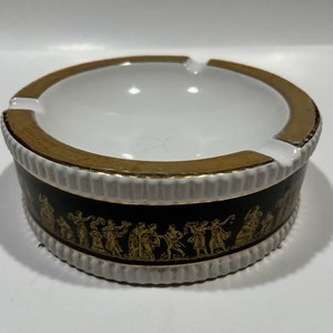 Op de afbeelding: Een witte asbak van keramiek met een gouden rand en een zwarte band met een gouden ontwerp van mensen die rond een boom dansen.
