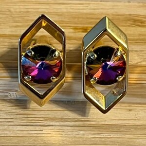 Könnte beinhalten: Ein Paar goldfarbene Ohrstecker. Jeder Ohrring hat einen großen, schillernden, runden Edelstein in den Farbtönen Lila, Rosa und Gold, der in einem geometrischen, offenen Rahmen eingefasst ist. Die Ohrringe werden auf einer Holzoberfläche präsentiert.