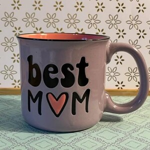May include: A lavender ceramic mug with the words "best MOM" in black lettering and a pink heart. The interior of the mug is peach-coloured, and the handle is the same lavender colour as the exterior. The mug is on a patterned surface.