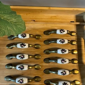 Peut inclure: Un ensemble de quincaillerie d'armoire, comprenant des poignées et des boutons de tiroir. Les poignées ont un centre en céramique blanche avec un motif floral et des extrémités en métal de couleur laiton. Les boutons ont un design assorti.