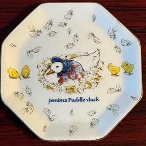 Peut inclure: Une assiette en céramique blanche avec une illustration de canard bleu et rouge. Le canard est entouré de poussins jaunes et de plumes blanches. Le texte "Jemima Puddle-duck" est imprimé sur l'assiette.