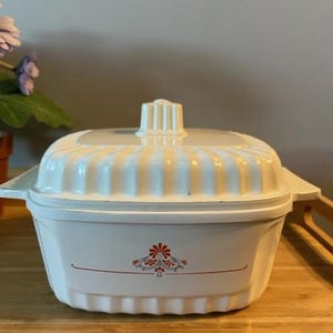 May include: White casserole dish with a lid. The dish has a red and orange floral design and a ribbed lid. The dish is sitting on a wooden surface.