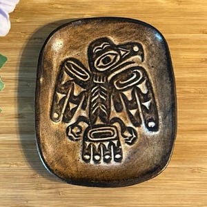 May include: A brown, rectangular ceramic dish featuring a raised, stylised eagle design. The eagle has outstretched wings and intricate details. The dish has a slightly textured surface and a dark brown colour.
