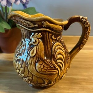 May include: A brown ceramic jug featuring a rooster design. The jug has a detailed relief pattern of a rooster, corn, and floral elements. The curved handle is decorated with floral accents. The jug is likely vintage and has a glossy finish.