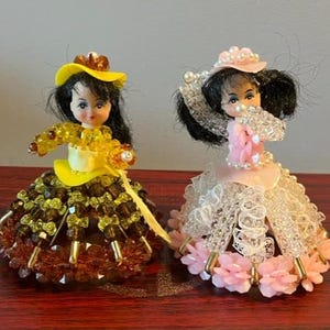 Könnte beinhalten: Zwei vintage Perlenpuppen mit Hüten und Kleidern. Die Puppe links trägt einen gelben Hut und ein braunes Kleid mit grünen Perlen. Die Puppe rechts trägt einen rosa Hut und ein weißes Kleid mit rosa Perlen.