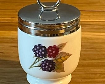 Könnte beinhalten: Weißer Eierbecher aus Keramik mit silbernem Deckel und Griff. Der Becher ist mit einem Brombeermotiv in Lila- und Rottönen mit grünen Blättern verziert. Der Rand und der Boden haben eine silberne Zierleiste. Der Eierbecher steht auf einer Holzoberfläche.