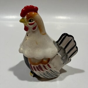 Può includere: Un set di saliera e pepe in ceramica a forma di pollo. Il pollo è bianco con strisce nere e rosse e una cresta rossa. Il pollo è in piedi su una superficie bianca.