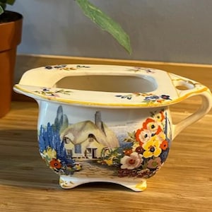 May include: Cream ceramic chamber pot with a handle and a rectangular opening. The pot features a painted landscape scene with a cottage, trees, and flowers in blue, yellow, and red. Floral accents decorate the rim.