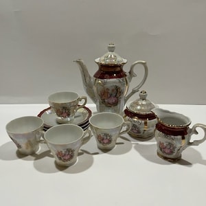 May include: A vintage porcelain tea set with a floral design. The set includes a teapot, creamer, sugar bowl, and four teacups and saucers. The teacups and saucers have a delicate floral pattern with gold accents. The teapot has a red and gold design with a floral pattern.