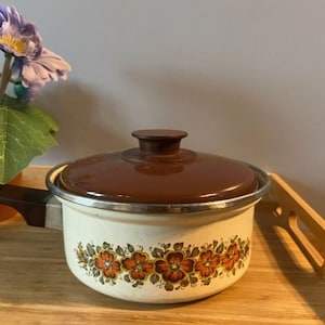 Pode incluir: Uma panela vintage creme com tampa e alça marrons. A panela é decorada com um padrão floral em tons de laranja e marrom. A panela está em uma bandeja de madeira.