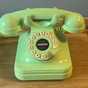 Può includere: Un telefono rotante verde menta in stile vintage con una cornetta e un cavo a spirale. Il telefono presenta un quadrante circolare con pulsanti numerici bianchi e le parole "FLASH REDIAL".