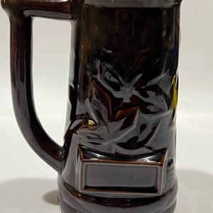 May include: A brown ceramic beer stein with a handle and a leaf design. The stein has a blank rectangular space on the front.