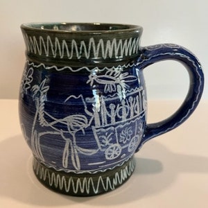 Pode incluir: Uma caneca de cerâmica azul com um desenho branco de uma carroça puxada por um cavalo e pessoas. A caneca tem uma borda e uma base verdes.