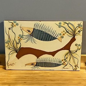 Op de afbeelding: Rechthoekige keramische tegel met twee gestileerde vissen in blauw en wit. De crèmekleurige achtergrond heeft bruine takken en groen gebladerte. De tegel heeft handtekeningen in de onderste hoeken. Een decoratief stuk.