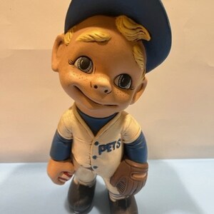 May include: A ceramic figurine of a baseball player wearing a blue hat with the word "PETS" on the front of the jersey. The figurine is holding a baseball bat and a glove.