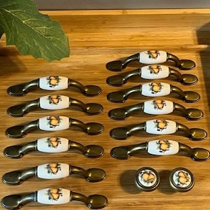 Puede incluir: Un conjunto de herrajes para armarios, que incluye tiradores y pomos. Los tiradores tienen una base de metal curvada de color bronce con un centro de cerámica blanca. Los centros de cerámica presentan un diseño de fruta. También se incluyen dos pomos a juego.