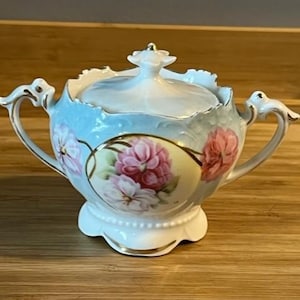 May include: A white porcelain sugar bowl with a lid, featuring a floral design with pink, white, and yellow flowers on a light blue background. The bowl has a gold trim and a scalloped edge.
