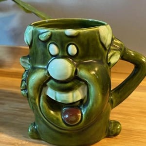May include: Green ceramic mug shaped like a cartoon face with a wide smile and a red nose. The mug has a handle on the right side.