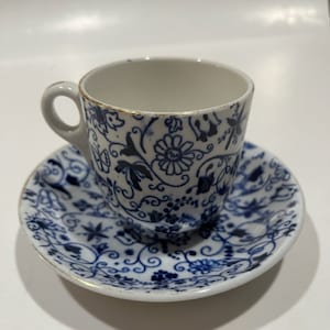 以下が含まれることがあります： 青い花柄の白いティーカップとソーサーのセット。カップには小さな取っ手と金の縁があります。ソーサーにも金の縁があります。このセットはおそらく磁器製です。