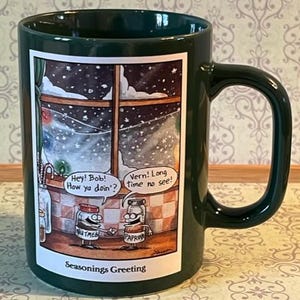 Könnte beinhalten: Dunkelgrüne Keramiktasse mit einer Cartoon-Illustration. Die Illustration zeigt zwei Gewürzgläser, Muskatnuss und Paprika, die sich vor einem verschneiten Fenster die Hände schütteln. Auf der Tasse steht "Seasonings Greeting."