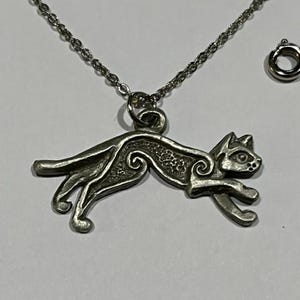 May include: A silver-toned cat pendant necklace. The pendant is a stylized cat in a running pose, with etched details. The necklace chain and clasp are also silver-toned. The cat pendant is approximately 1.5 inches long.