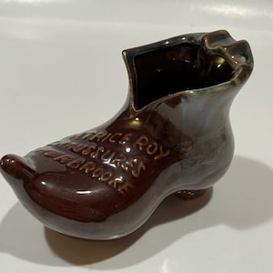 May include: A brown ceramic shoe-shaped vase with the text "Alice Roy, chaussures, BROOKE" embossed on the side. The vase has a glossy finish and a small opening at the top. The shoe has a rounded toe and a small heel.