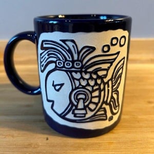 May include: A navy blue ceramic mug with a white, stylized fish design. The mug features a curved handle and a detailed illustration of a fish with intricate patterns and shapes. The design is reminiscent of ancient art.