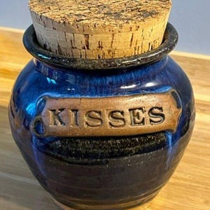 May include: A small, blue ceramic jar with a cork stopper. The jar has a brown label with the word "KISSES" embossed on it. The jar is round with a dark blue glaze and a black base.