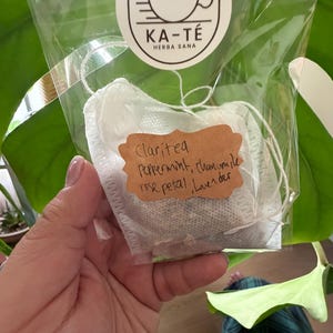 May include: A clear plastic bag containing a tea bag with a brown label. The label reads "Claritea" and lists ingredients: peppermint, chamomile, rose petal, and lavender. The bag also has a round sticker with the text "KA-TÉ HERBA SANA" and a teacup logo.