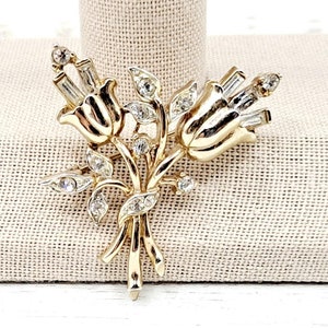 May include: A gold-toned brooch featuring two stylized flower buds with clear rhinestone accents. The brooch has a delicate, vintage aesthetic.