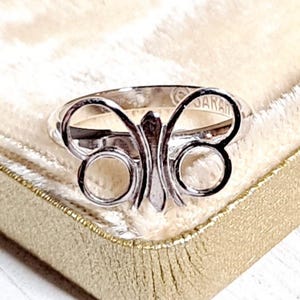 May include: A silver-colored ring with a butterfly design. The ring features a central vertical element and two rounded sections on each side, forming the butterfly's wings. The word "JAPAN" is stamped on the ring.
