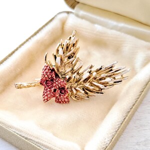 May include: A gold-toned brooch in the shape of a wheat stalk with a cluster of red jewels. The brooch is displayed in a beige velvet-lined box with a gold-colored trim. The brooch has a textured surface and is likely vintage.