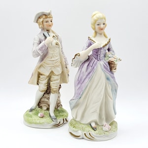 May include: A pair of porcelain figurines depicting a man and a woman in 18th-century attire. The man is wearing a light purple jacket, white shirt, and brown pants. He is holding a fan in his left hand. The woman is wearing a lavender dress with a white underskirt. She is holding a basket of flowers in her right hand.