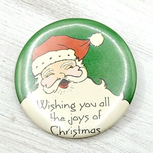 May include: A green and white Christmas button with a cartoon image of Santa Claus. The button reads "Wishing you all the joys of Christmas".