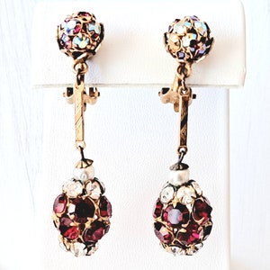May include: A pair of dangle earrings with a gold tone finish. The top of each earring features a round cluster of red, blue, and clear rhinestones. The bottom of each earring features a round cluster of red rhinestones with a pearl accent.