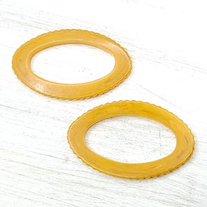 May include: Two oval-shaped, amber-colored bangles with a scalloped edge are displayed on a white, textured surface. The bangles are made of a translucent material, and the light reflects off their smooth surfaces.