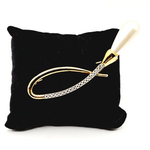 May include: A gold-tone brooch with a pearl-shaped accent and a row of clear rhinestones. The brooch is displayed on a black velvet cushion.