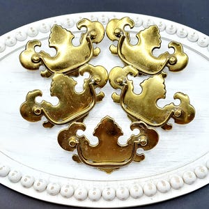 May include: Six antique brass drawer pulls with a decorative design. The pulls are arranged on a white oval tray with a beaded edge. The drawer pulls have a curved shape with ornate details, perfect for furniture restoration projects.