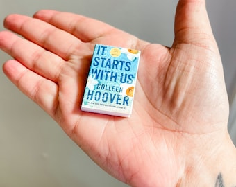 May include: A paperback book titled "It Starts With Us" by Colleen Hoover. The cover features a blue and white floral design with a yellow flower.