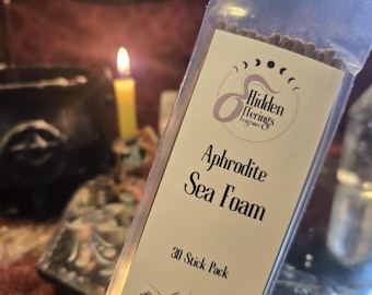 Op de afbeelding: Een transparante zak met wierookstokjes. Het etiket leest "Hidden Offerings Fragrance Co." met de geur "Aphrodite Sea Foam". Het etiket vermeldt ook "30 Stick Pack" en heeft een halve maan en een zonnestraalontwerp. Een brandende kaars is zichtbaar op de achtergrond.