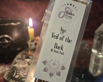 May include: A clear plastic package containing incense sticks. The label reads "Nyx Veil of the Dark 10 Stick Pack" with a crescent moon and sunburst design. The background includes a lit candle and a cauldron.