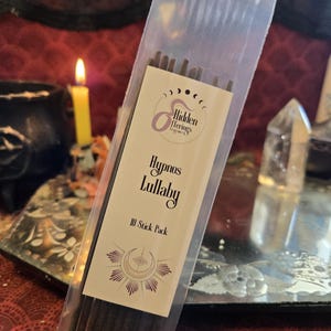 May include: A clear plastic package containing incense sticks. The label reads "Hidden Terrings" and "Hypnos Lullaby, 10 Stick Pack." The label features a crescent moon and sunburst design. A lit yellow candle and a crystal are visible in the background.