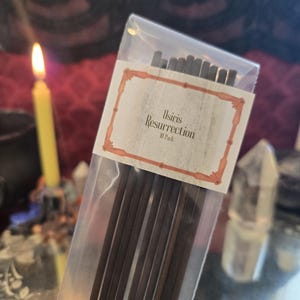 May include: A clear plastic package containing dark brown incense sticks. The label reads "Osiris Resurrection" in a decorative font. A lit yellow candle and a crystal are visible in the background.