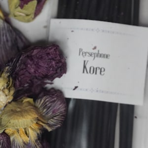 May include: Dried purple and yellow flower petals with a white label that reads "Persephone Kore". Black sticks are visible in the background. The image is shot from above.