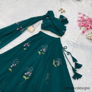 May include: A teal two-piece outfit featuring a crop top, a long skirt, and a matching scarf. The skirt and scarf are adorned with a floral pattern in pink and yellow. The outfit includes a decorative belt and tassel details.