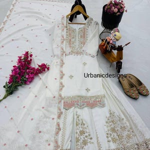 May include: White embroidered salwar kameez set with a matching dupatta. The set features intricate embroidery with pink and gold accents. The outfit is displayed on a hanger, with a bouquet of pink flowers and a pair of embellished shoes nearby. The text "Urban designs" is visible.