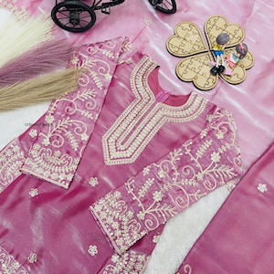 May include: A magenta-colored embroidered outfit with intricate gold-colored floral patterns. The outfit includes a long-sleeved top and a matching bottom. The top features a detailed neckline and sleeve cuffs. The outfit is displayed on a pink, textured surface.