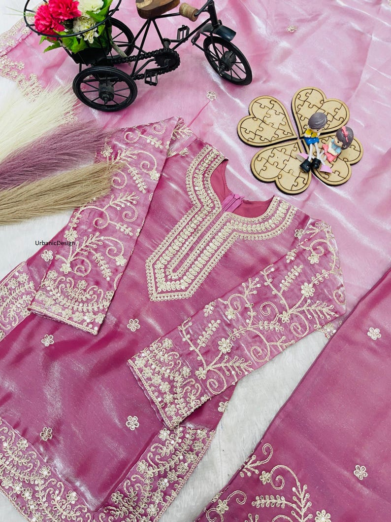 May include: A magenta-colored embroidered outfit with intricate gold-colored floral patterns. The outfit includes a long-sleeved top and a matching bottom. The top features a detailed neckline and sleeve cuffs. The outfit is displayed on a pink, textured surface.