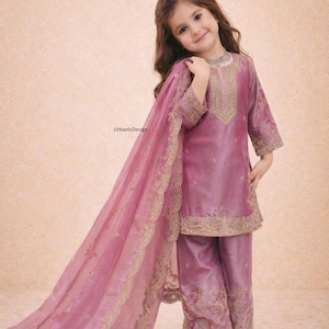 May include: A young girl wearing a mauve-colored traditional outfit. The ensemble includes a tunic, pants, and a flowing scarf, all adorned with gold embroidery. The outfit is made of a shiny fabric. The girl is smiling.