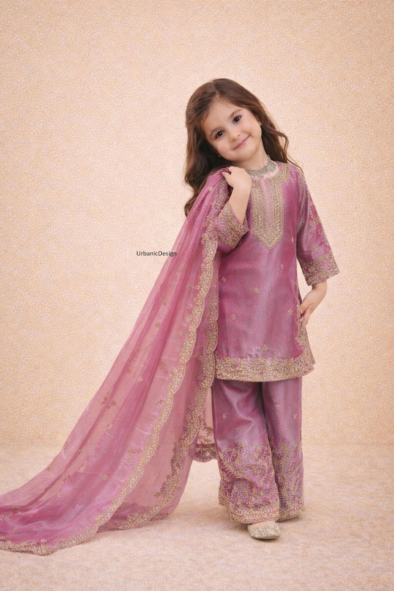 May include: A young girl wearing a mauve-colored traditional outfit. The ensemble includes a tunic, pants, and a flowing scarf, all adorned with gold embroidery. The outfit is made of a shiny fabric. The girl is smiling.